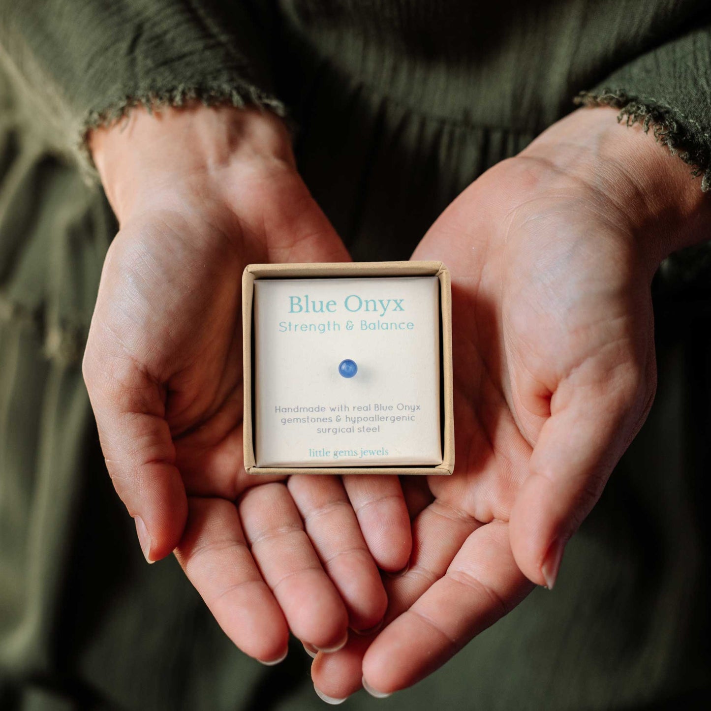 Woman wearing dark green dress holding gift box in hands containing tiny Blue Onyx gemstone stud earring.