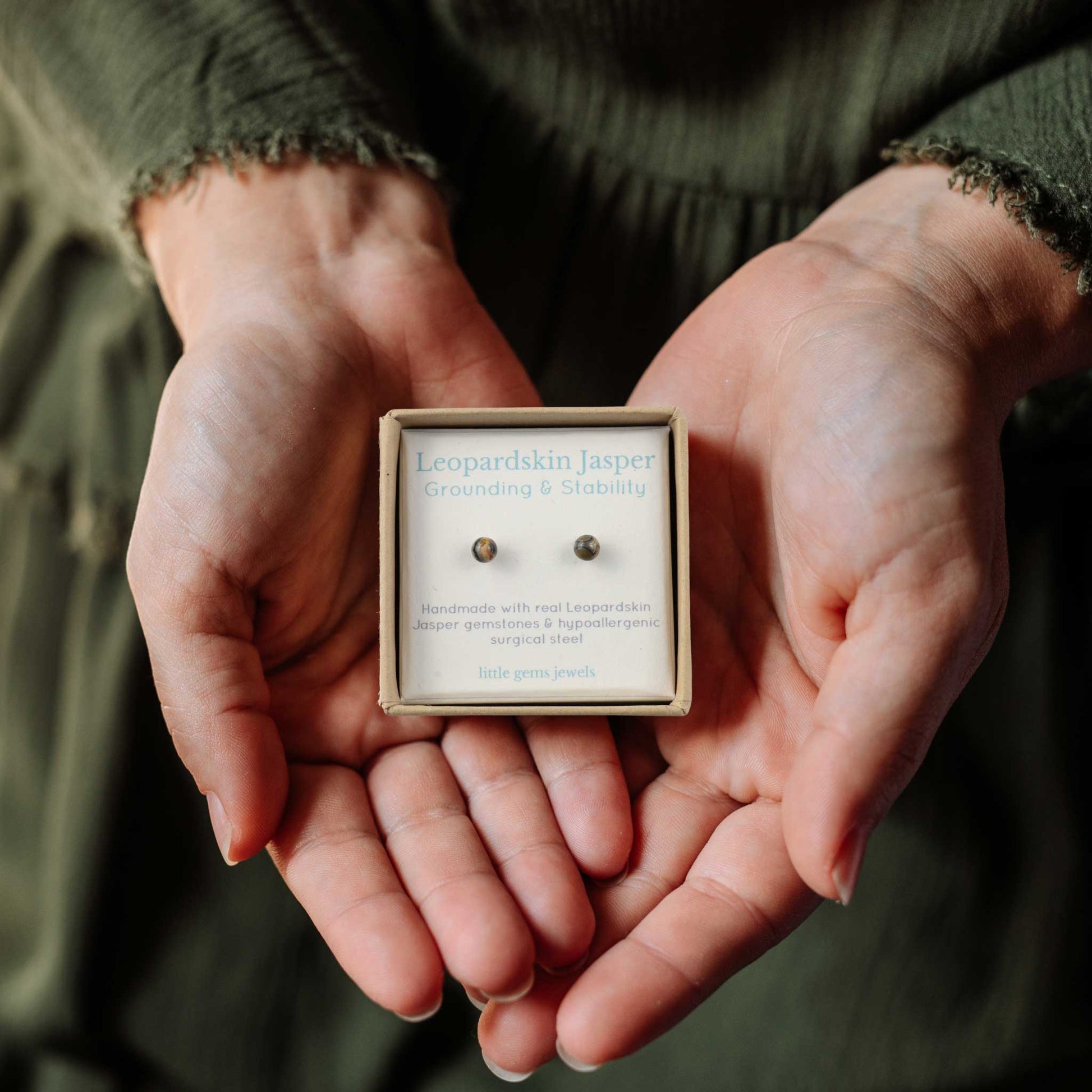 Hands holding small square eco friendly gift box containing tiny Leopardskin Jasper stud earrings.