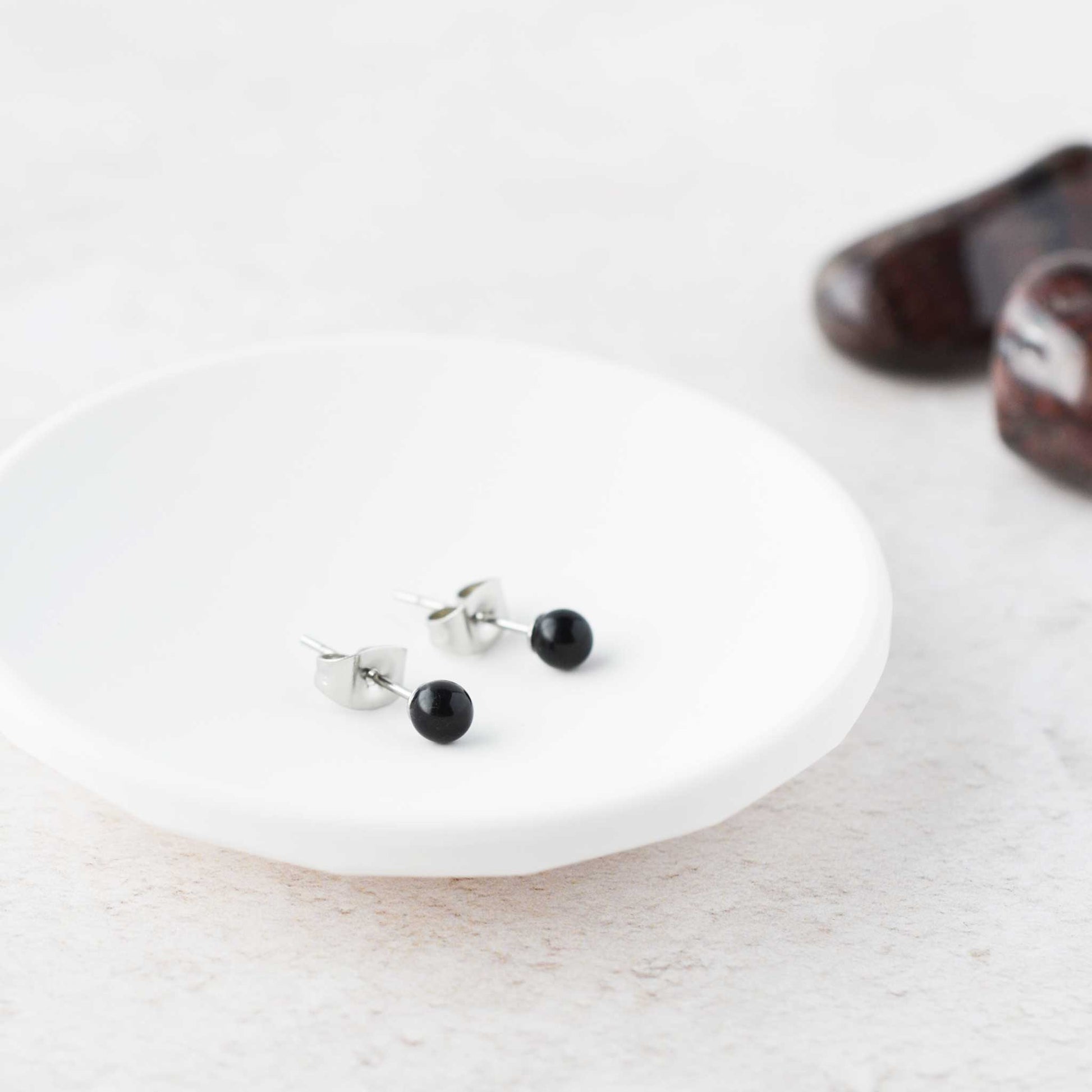 Tiny round black Onyx gemstone stud earrings lying in white trinket dish.