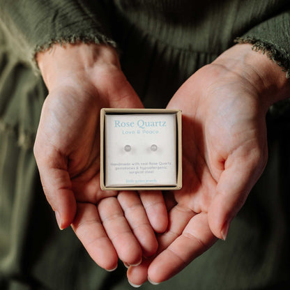 Hands holding small square eco friendly gift box containing tiny Rose Quartz stud earrngs.