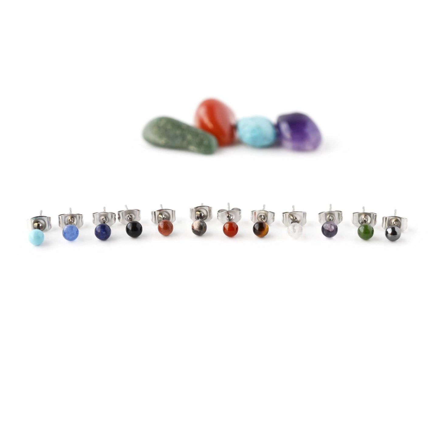 Twelve different coloured tiny single gemstone stud earrings in front of crystals on white background.