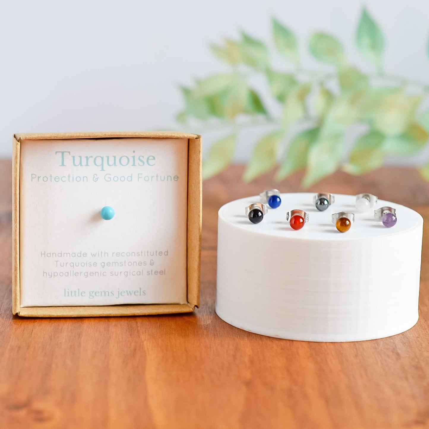 Turquoise single stud earring in box next to seven different coloured single stud earrings on white round plinth