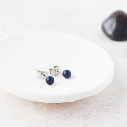 Tiny round dark blue gemstone stud earrings laying in white trinket dish