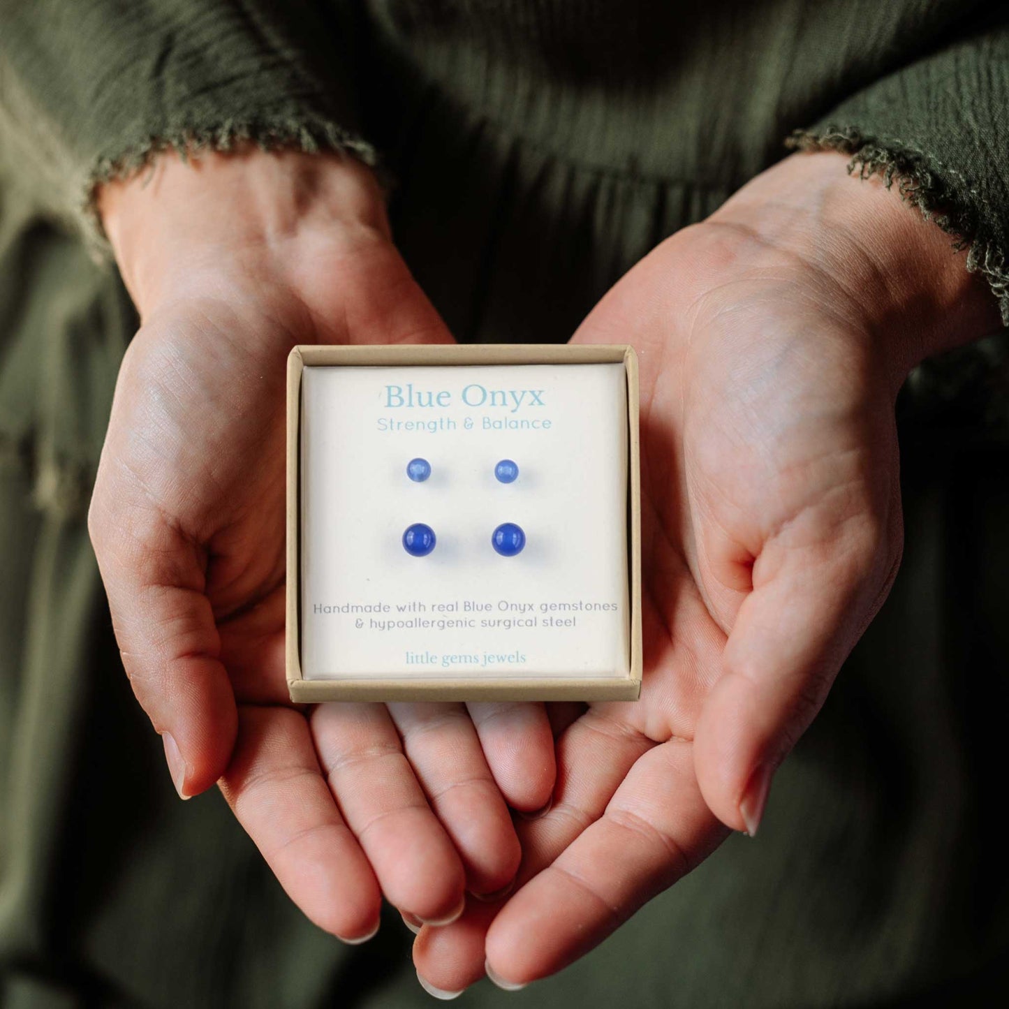 Hands holding a small box with Blue Onyx stud earrings inside on a dark background