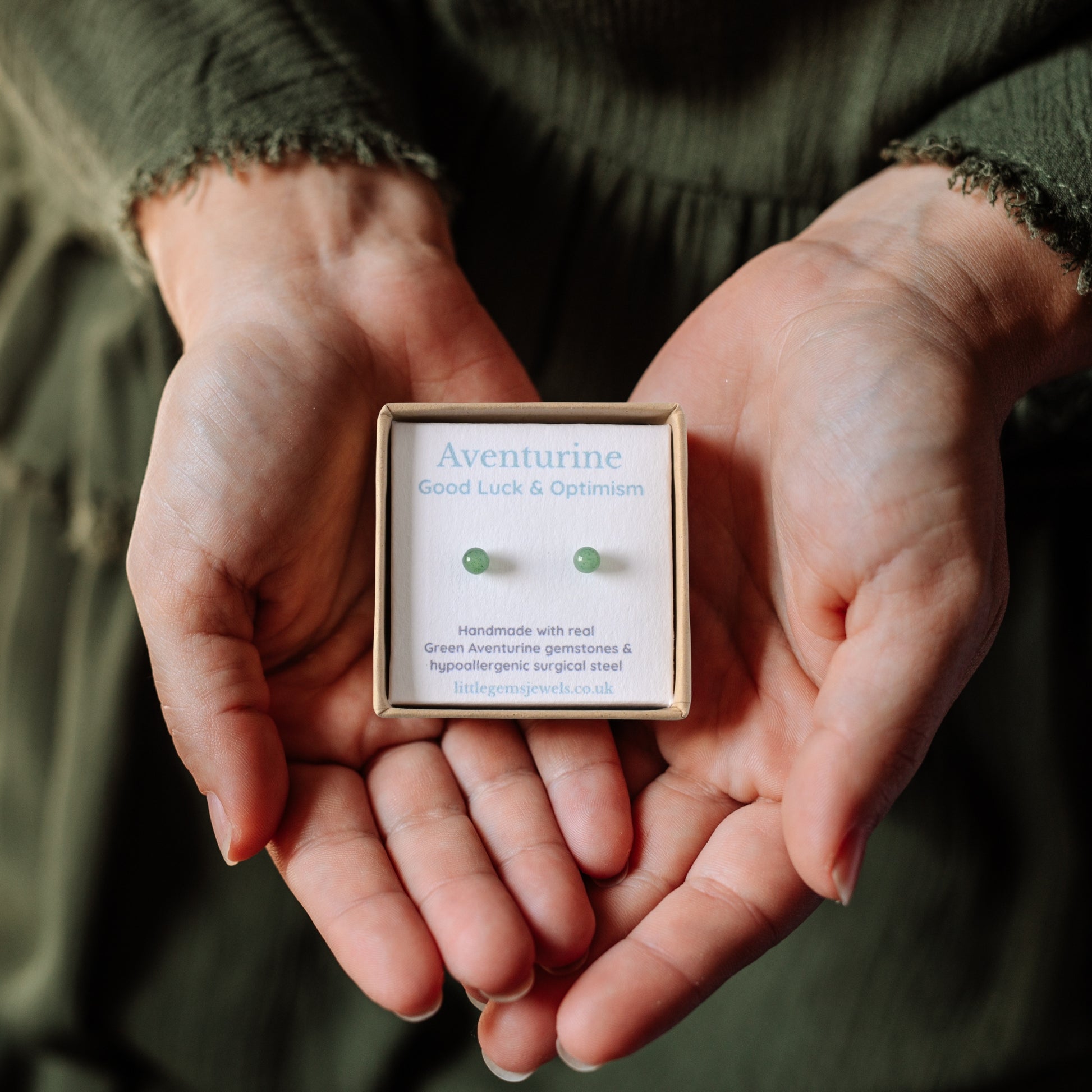 Hands holding a box with green aventurine earrings inside against a dark background