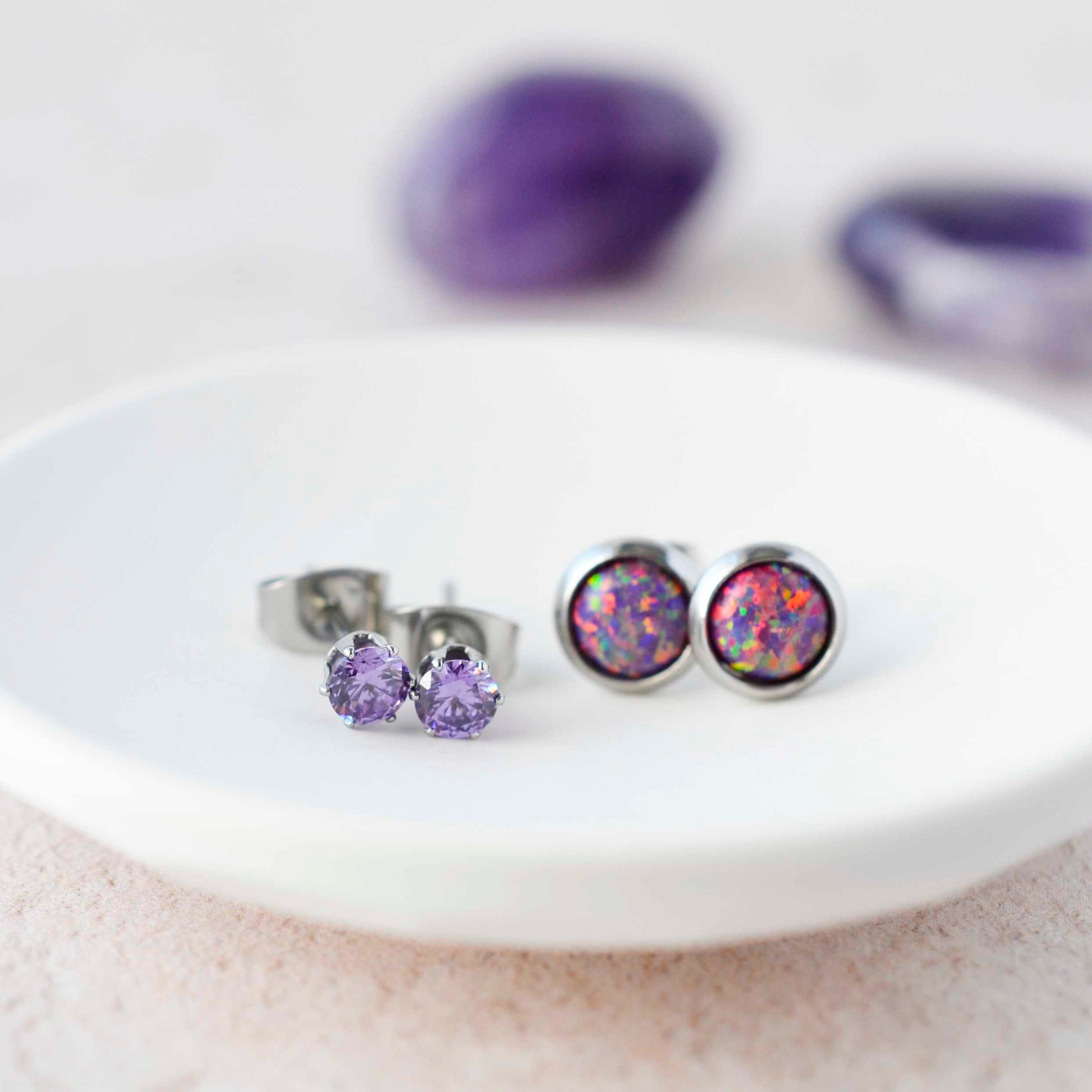 Purple CZ studs and purple lab Opal stud earrings sitting in a small white trinket dish.
