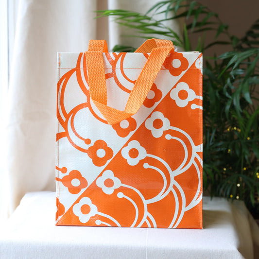 Orange and white patterned bag with matching handles on a white surface with a plant in the background.