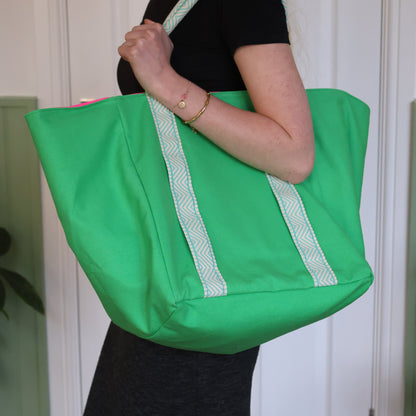 Person holding a green tote bag with white trim against a white wall.