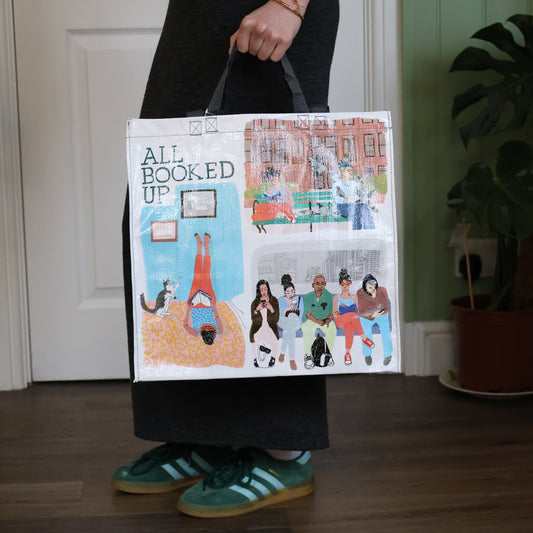 Person holding a tote bag with 'All Booked Up' design, standing indoors.