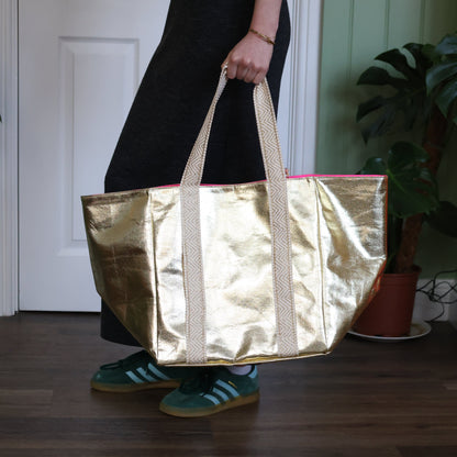 Person holding a gold metallic tote bag indoors with plants in the background