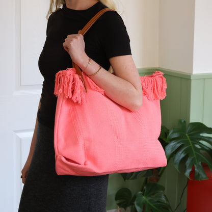 Person holding a pink tote bag with tassels in a room with a plant and red box.