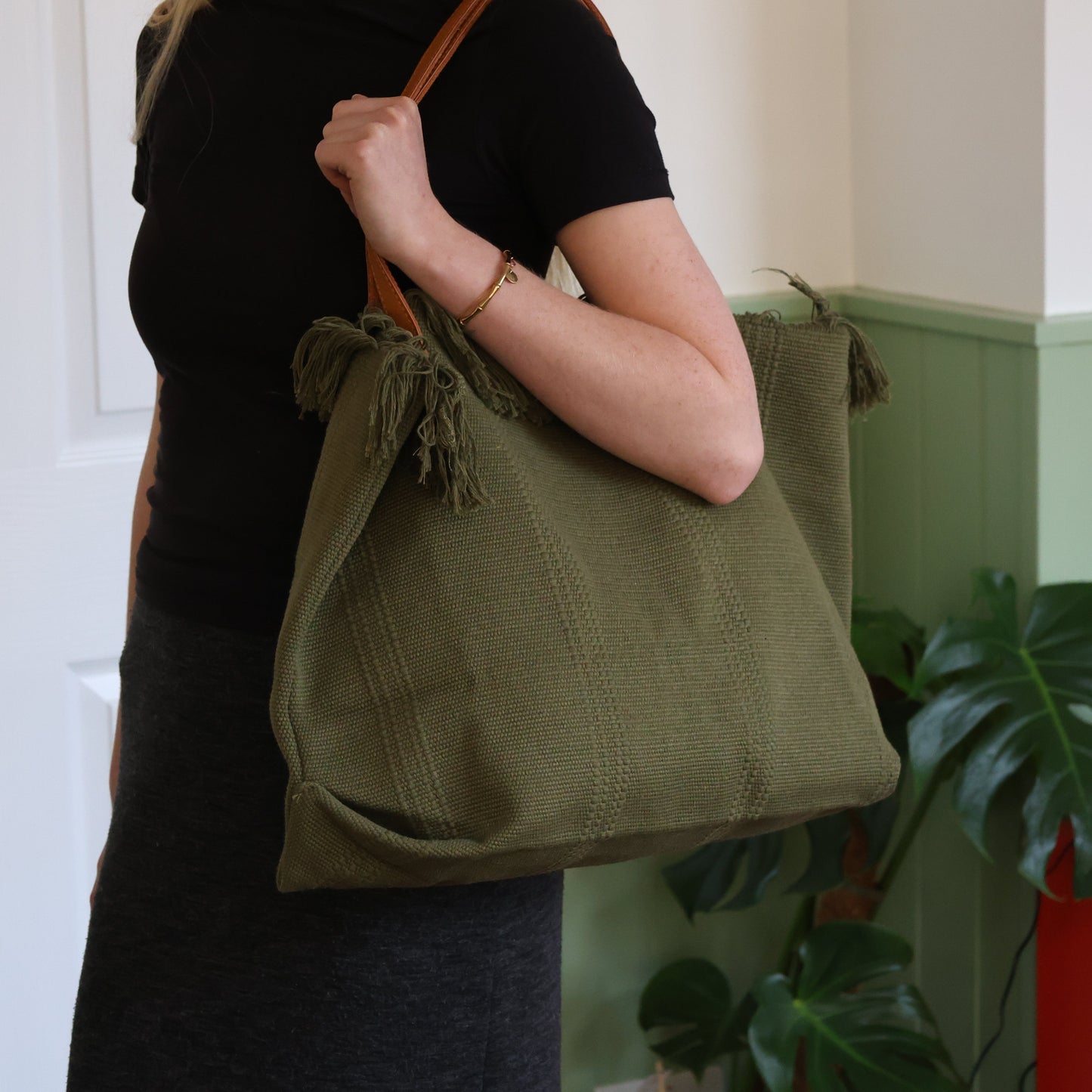Person holding a green tote bag with tassels against a light-colored wall.