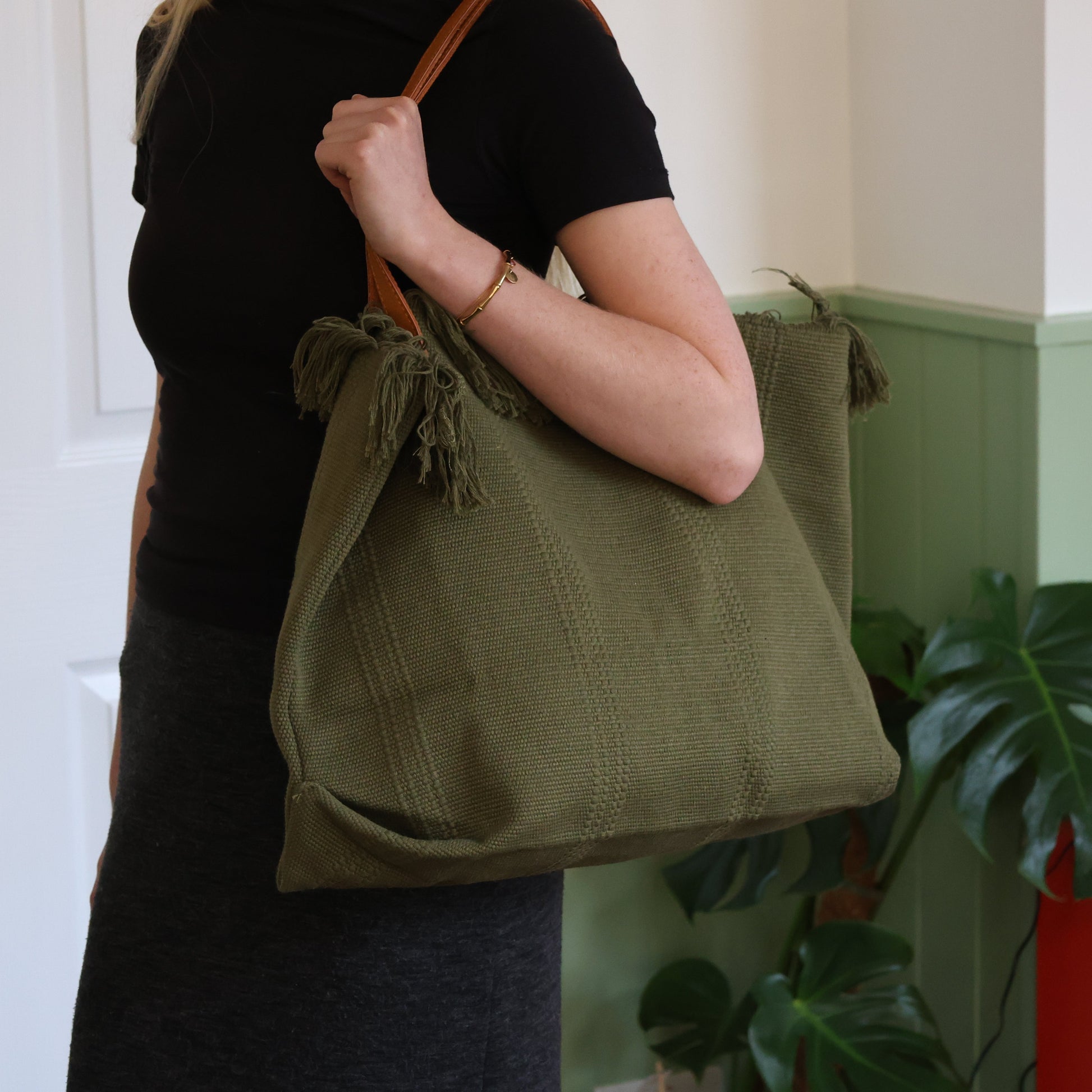 Person holding a green tote bag with tassels against a light-colored wall.