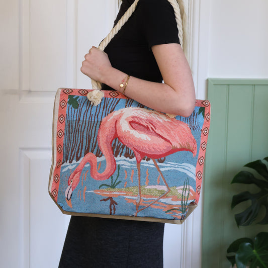 Person holding a tote bag with a flamingo design against a white wall.