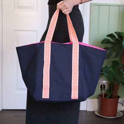 Person holding a navy blue tote bag with pink straps in a room with plants.