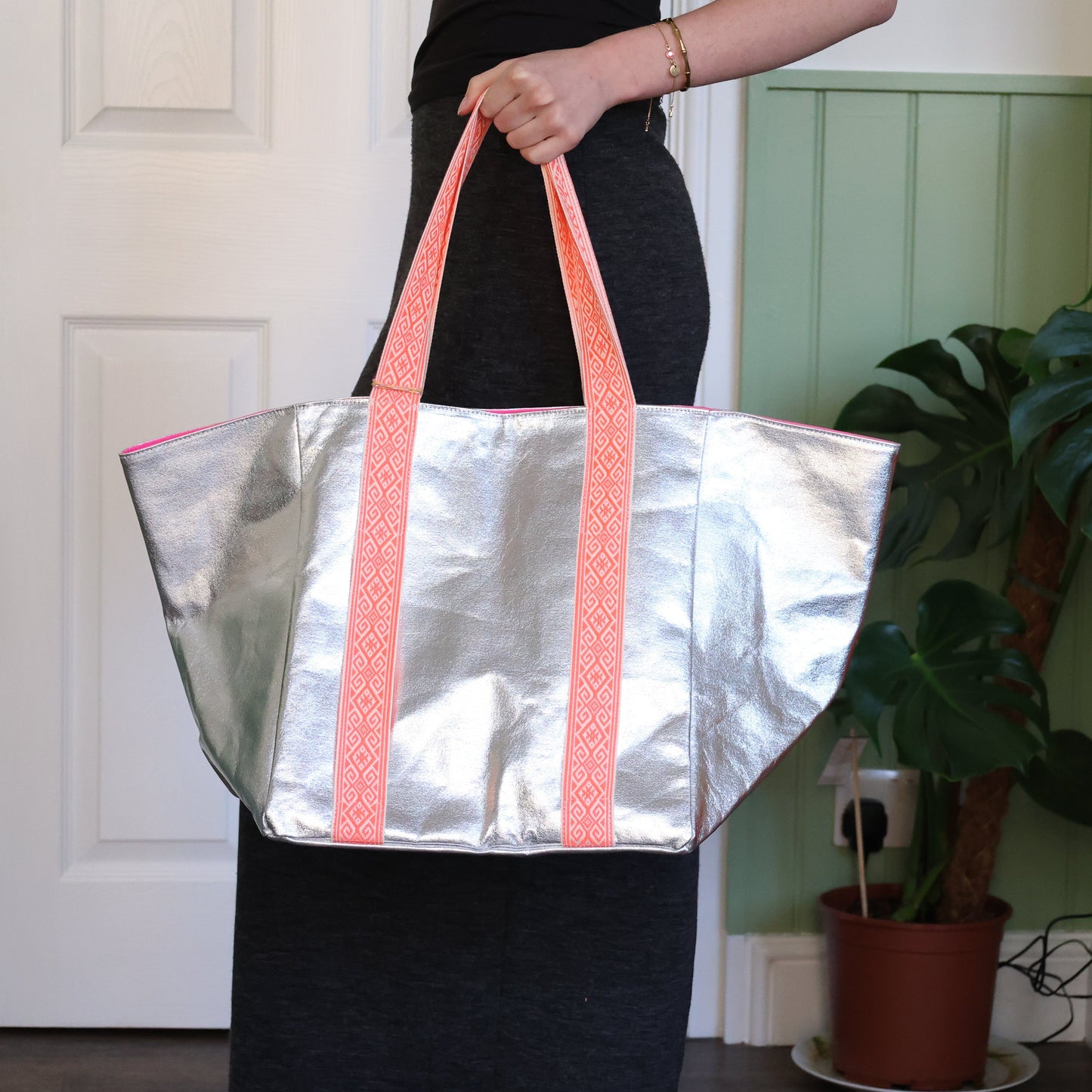 Person holding a silver tote bag with pink handles in a room with a plant.