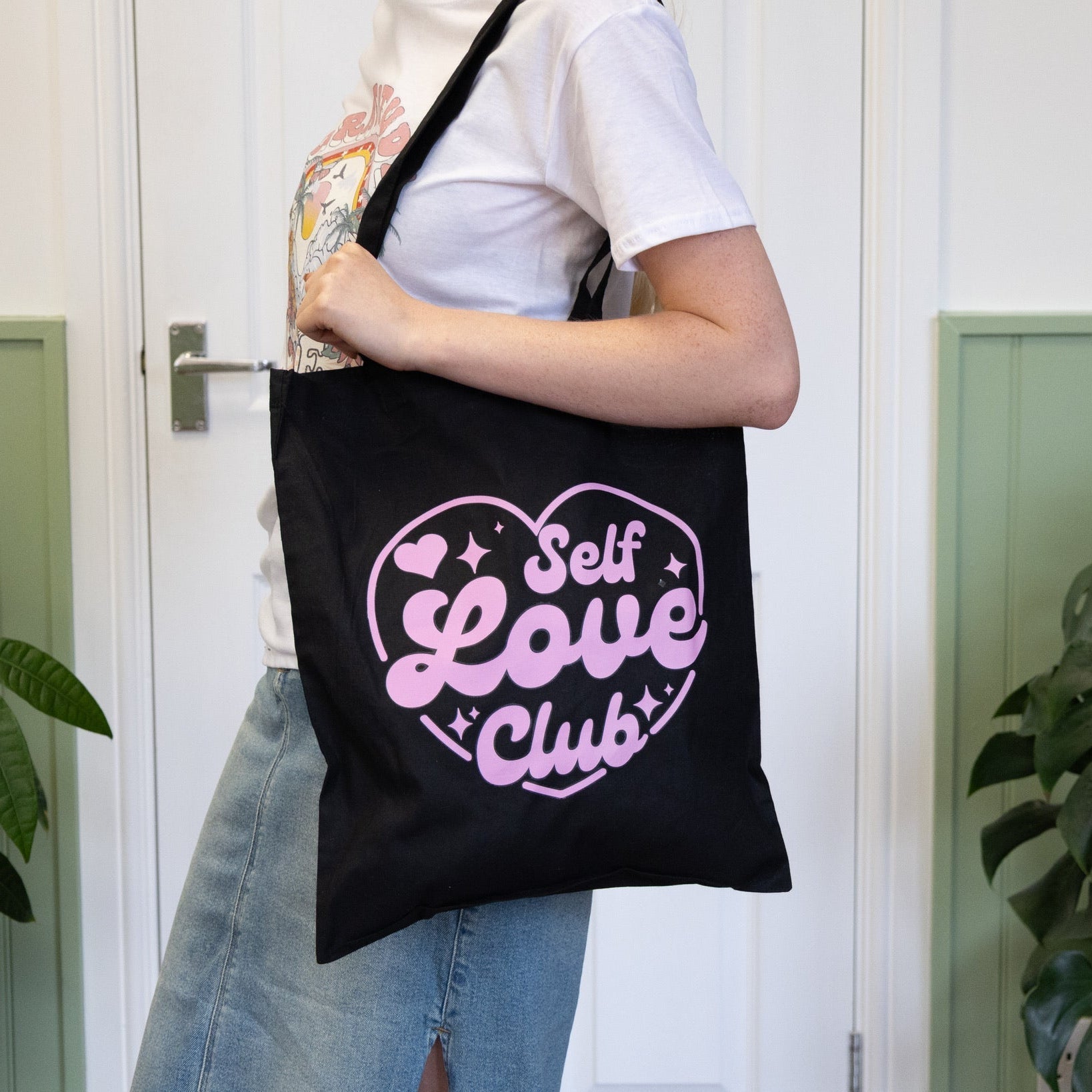 Person holding a black tote bag with 'Self Love Club' text in pink.