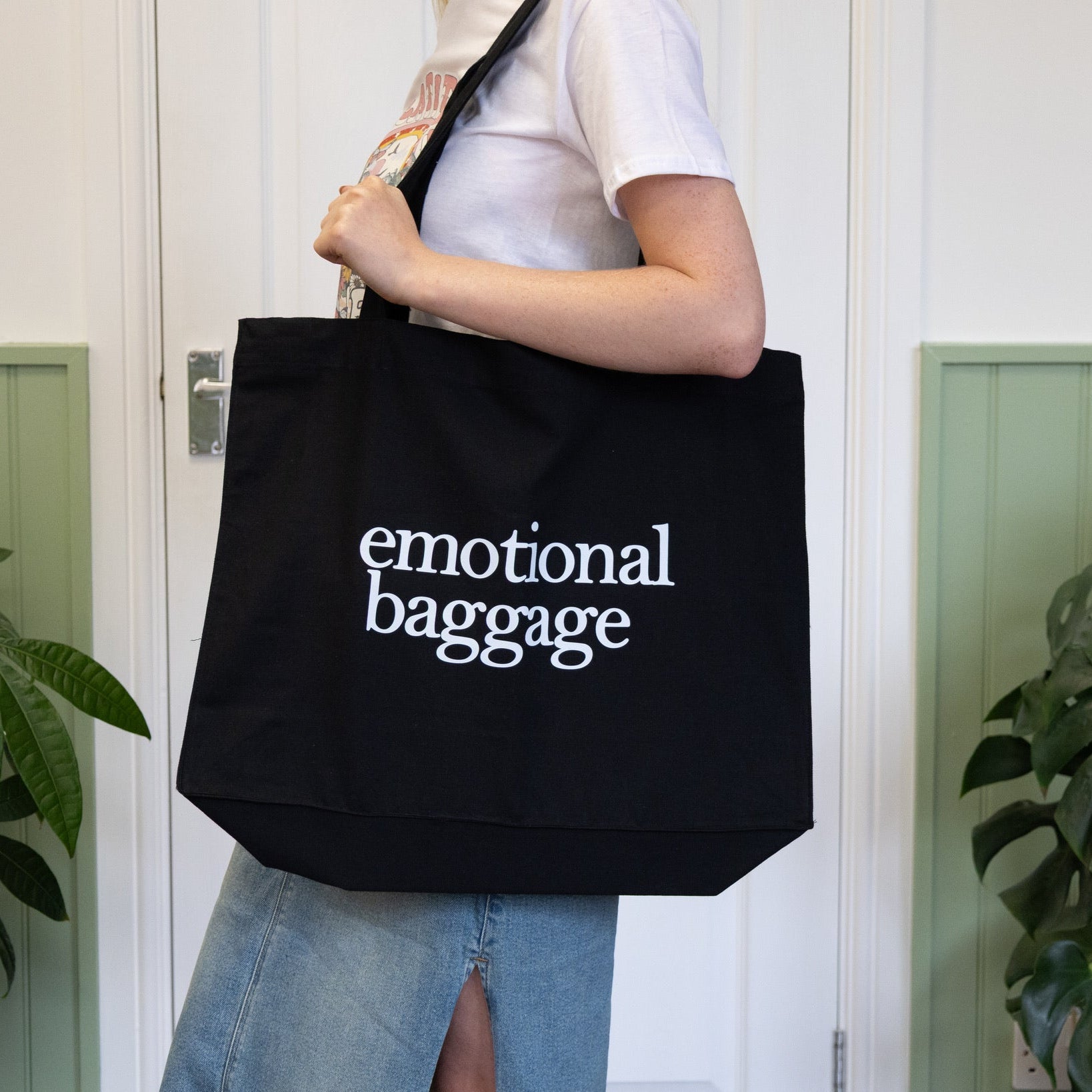 Person holding a black tote bag with 'emotional baggage' text in a room with plants.