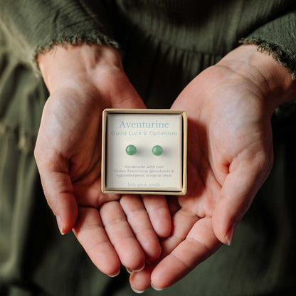 Hands holding a small square eco friendly gift box containing a pair of green aventurine stud earrings on a gemstone meaning card for good luck and optimism.