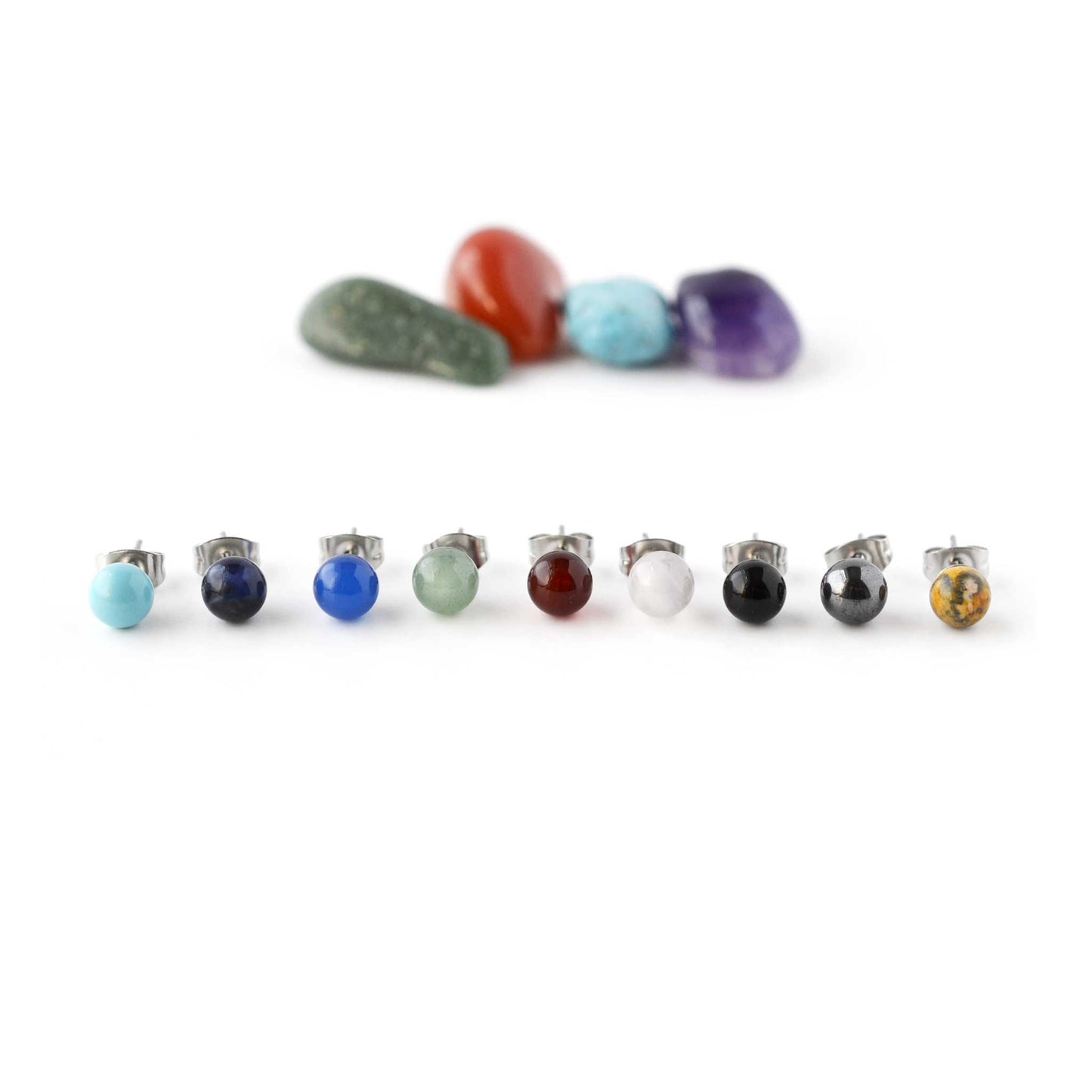 Nine different coloured gemstone single stud earrings in front of crystals on white background.