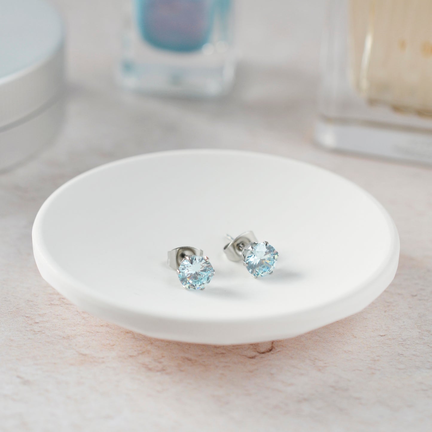 Pale blue stone stud earrings in white trinket dish against soft focus background.