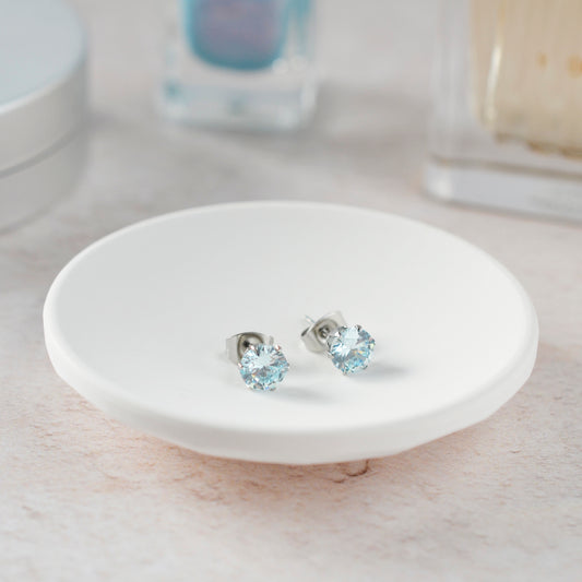 Pale blue stone stud earrings in white trinket dish against soft focus background.