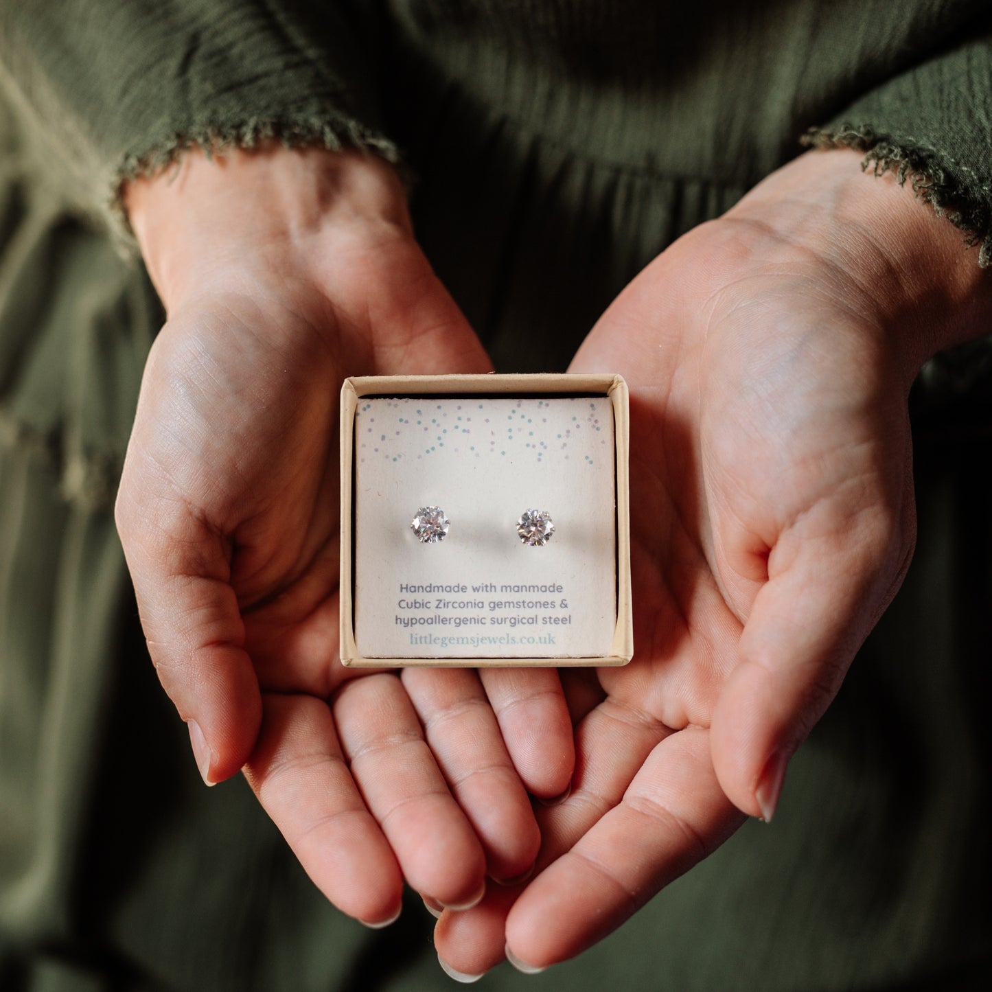Hands holding a box of CZ gemstone earrings with a blurred green background