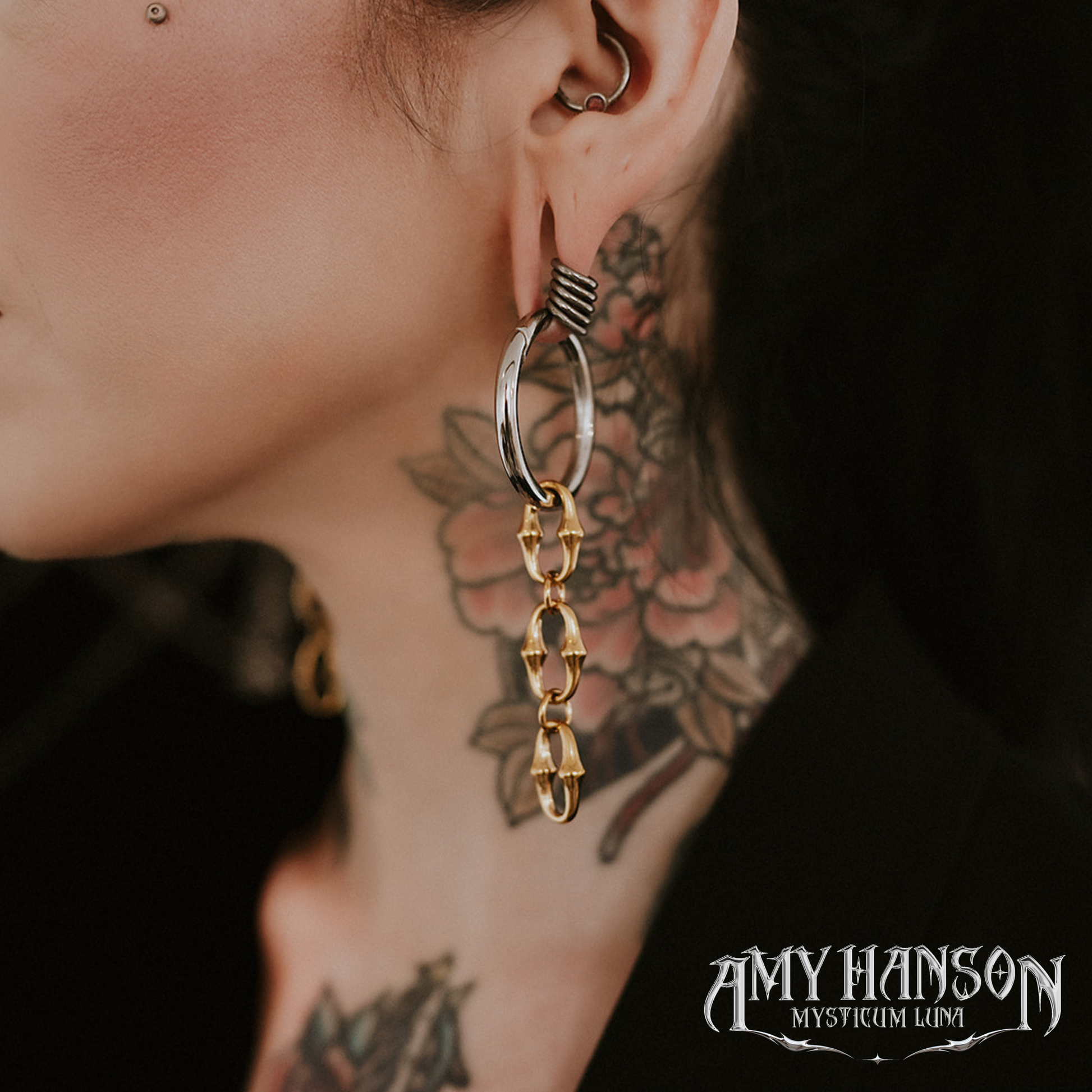 Close-up of an ear with a large hoop earring and tattoos on the neck, with 'Amy Hanson Mysticum Lumi' branding.