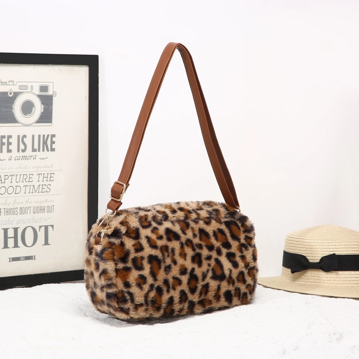 Leopard print handbag with brown strap on a white surface next to a framed picture and straw hat.