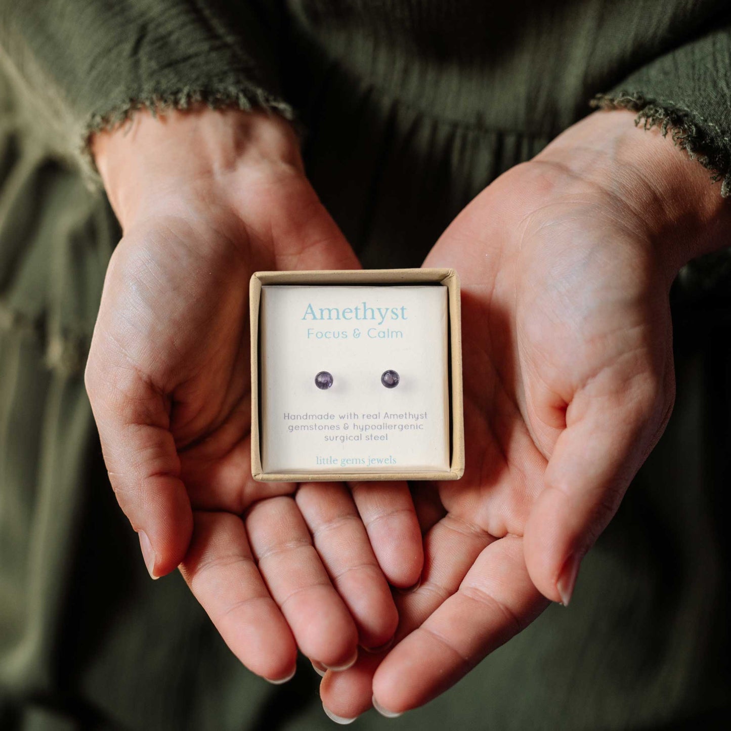 Hands holding a small square eco-friendly gift box containing tiny Amethyst stud earrings.