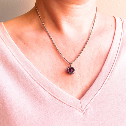 A close-up of a person's neck and chest wearing a silver-toned chain necklace with a round purple gemstone pendant, paired with a light pink V-neck top.