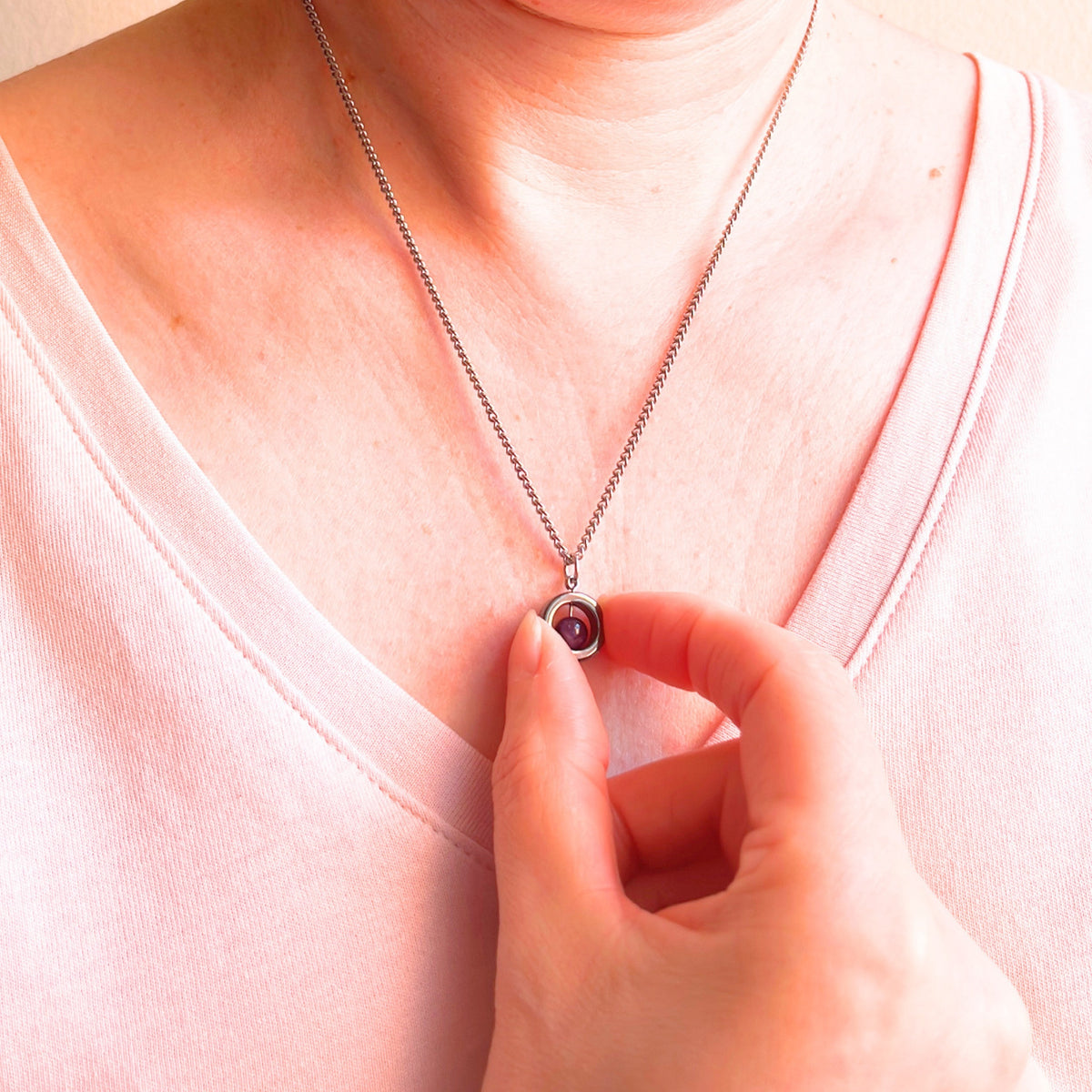 A close-up of a person wearing a delicate silver chain necklace with a small gemstone pendant, while wearing a light pink V-neck top.