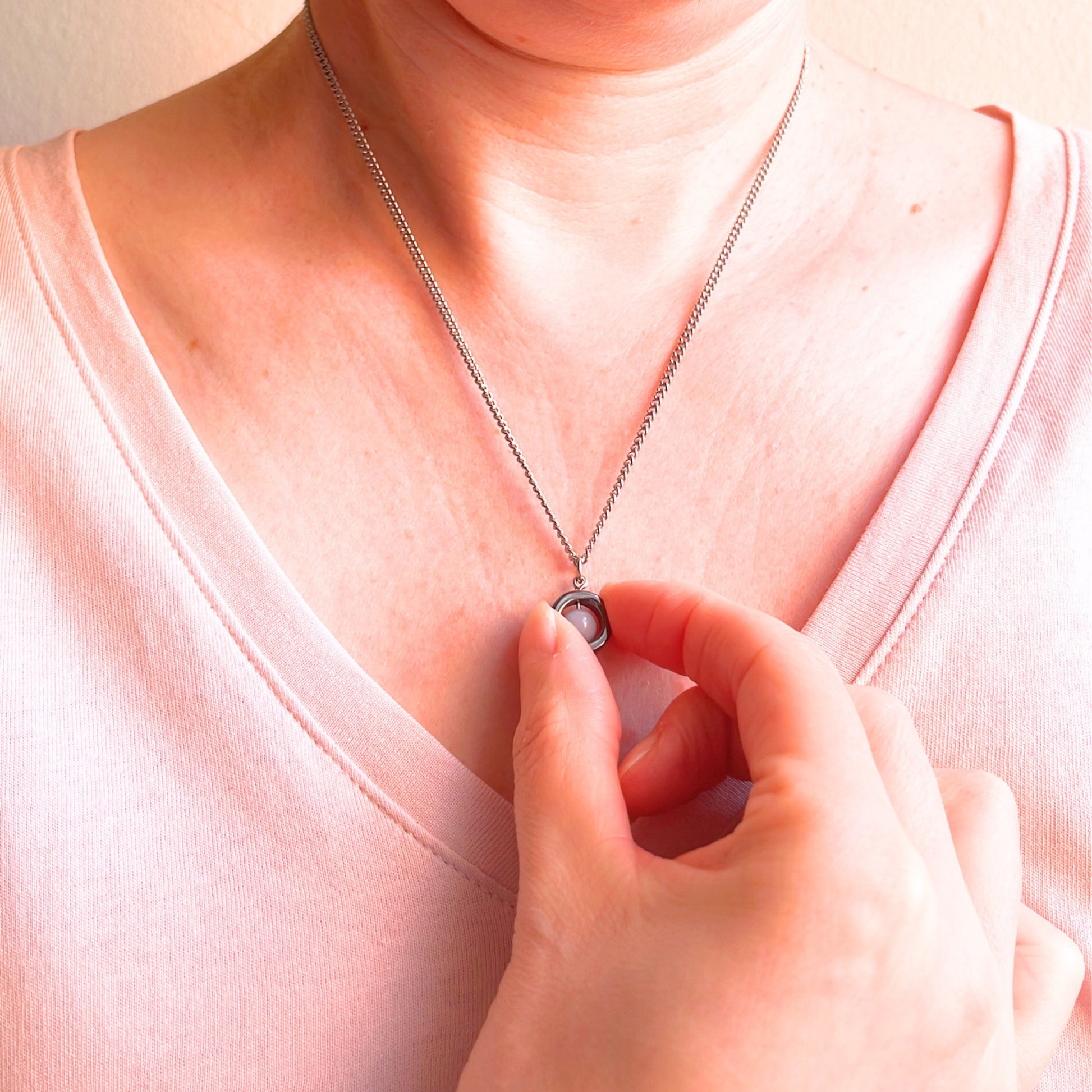 A close-up of a person wearing a delicate silver chain necklace with a small circular pendant, touching the pendant with their hand while wearing a light pink top.