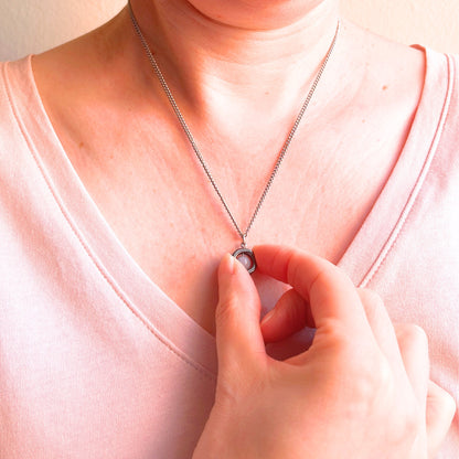 A close-up of a person wearing a delicate silver chain necklace with a small circular pendant, touching the pendant with their hand while wearing a light pink top.