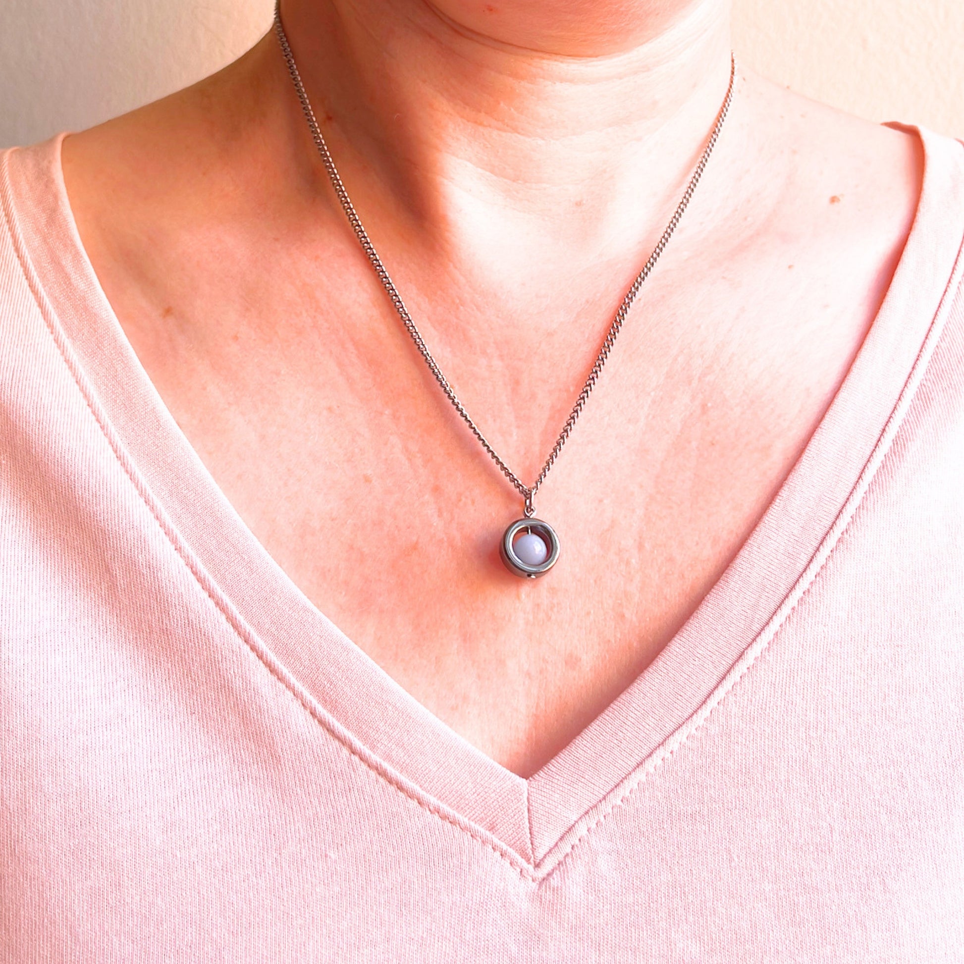 A close-up of a person's neck and chest wearing a silver chain necklace with a circular pendant featuring a light blue stone, paired with a light pink V-neck top.