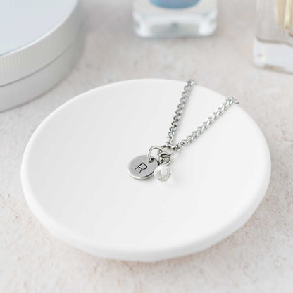 Tiny necklace with round disc pendant stamped with letter R and Rock Crystal gemstone charm laying in a white trinket dish.