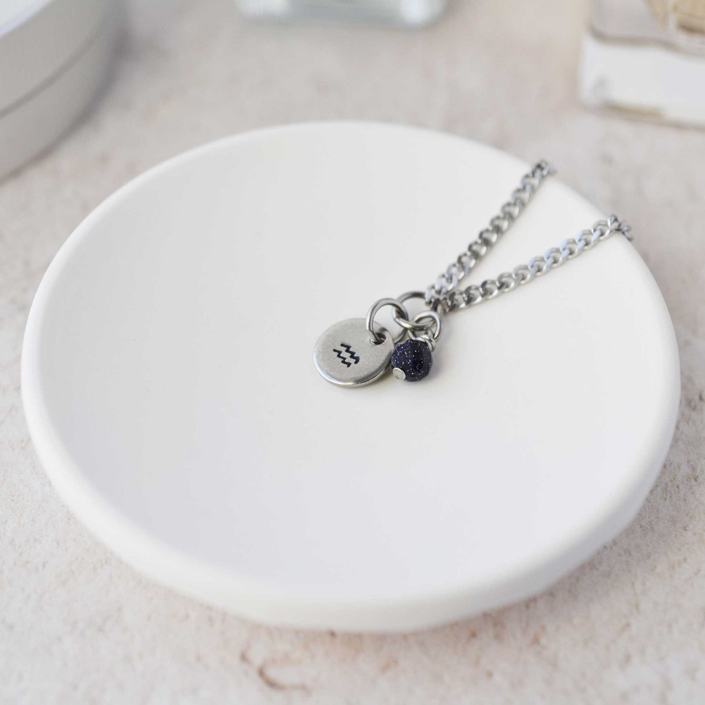 Tiny Aquarius zodiac sign disc pendant necklace with Blue Goldstone charm laying on white trinket dish