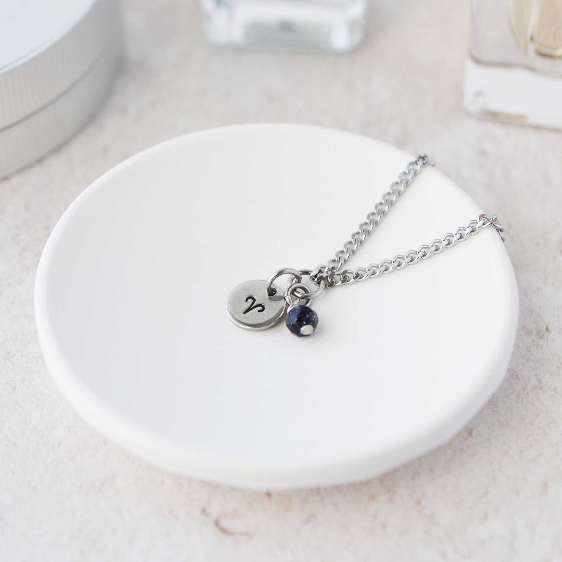 Tiny Aries zodiac sign disc pendant necklace with Blue Goldstone charm laying on white trinket dish