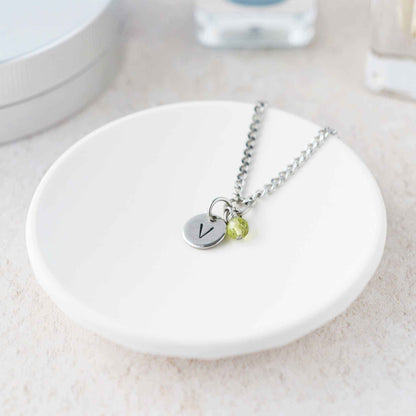 Tiny necklace with round disc pendant stamped with letter V and Peridot gemstone charm laying in a white trinket dish.