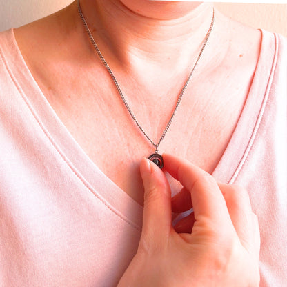 A close-up of a person wearing a delicate silver chain necklace with a small circular pendant, touching the pendant with a hand while wearing a light pink V-neck top.
