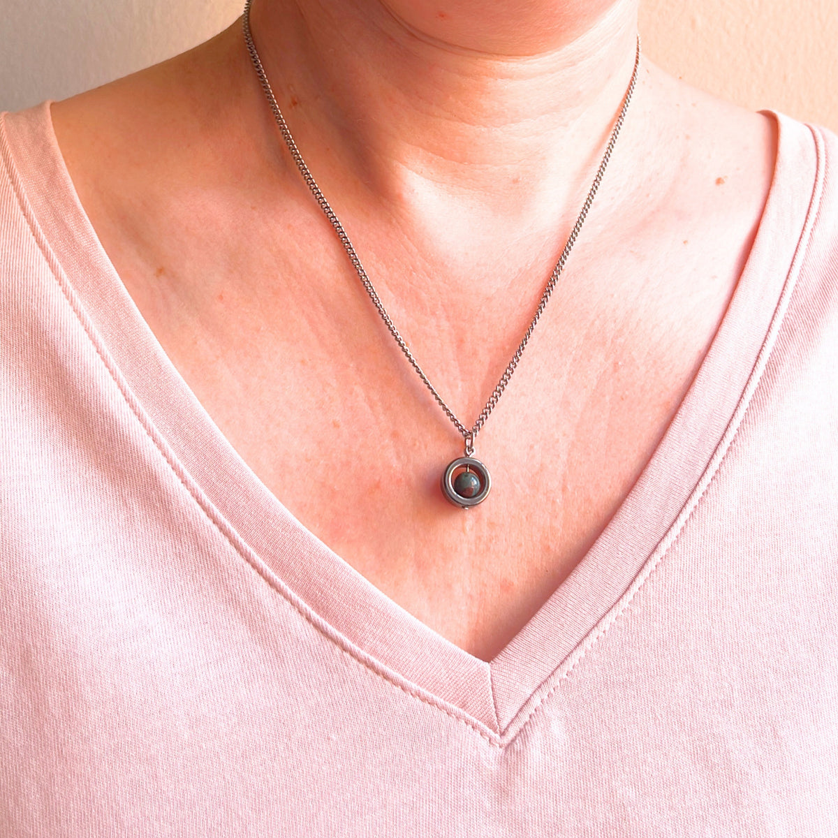 A close-up of a person's neck and chest wearing a silver-toned chain necklace with a circular pendant featuring a darkgreen gemstone, paired with a light pink V-neck top.