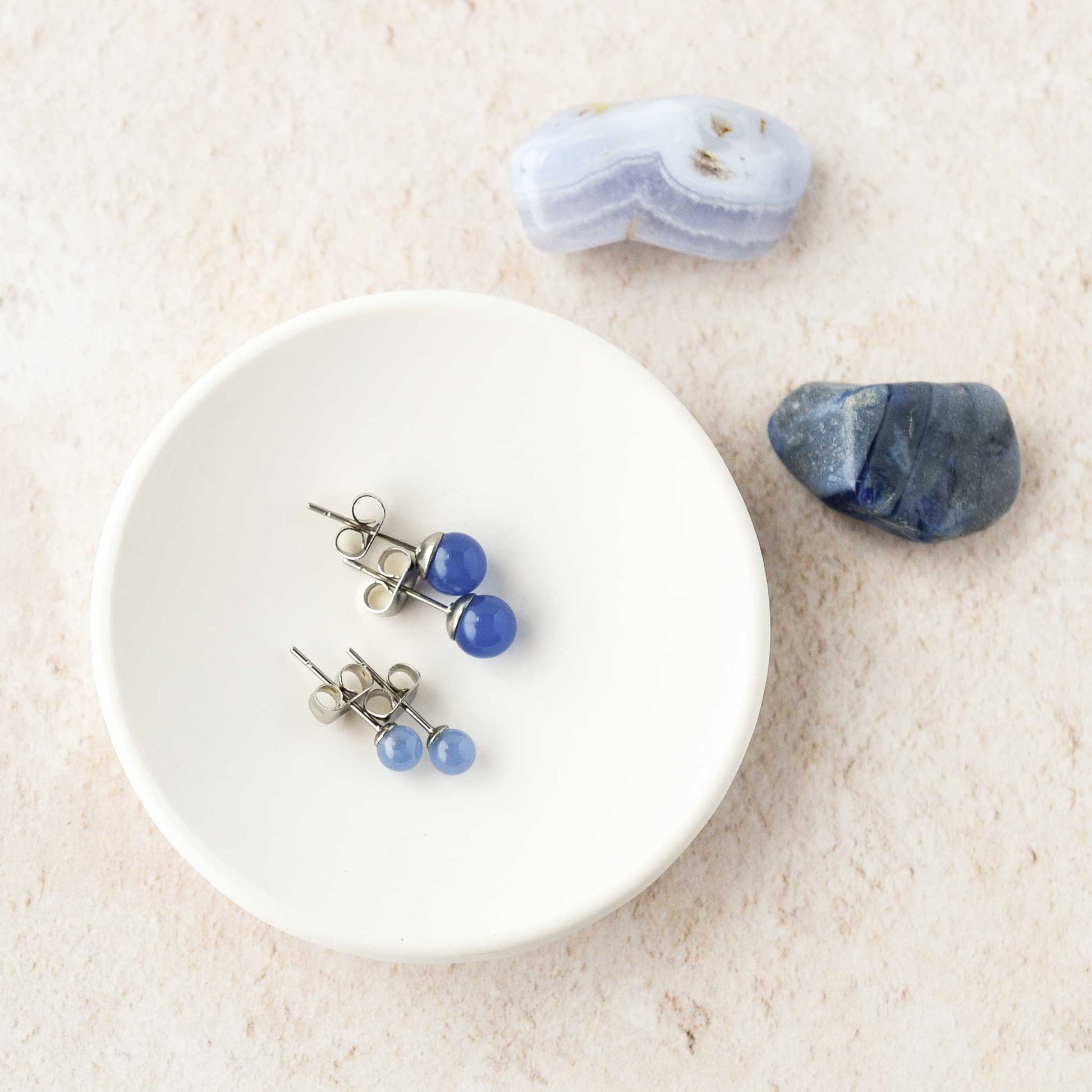 Two pairs of blue stone stud earrings on a white dish with stones on a beige background