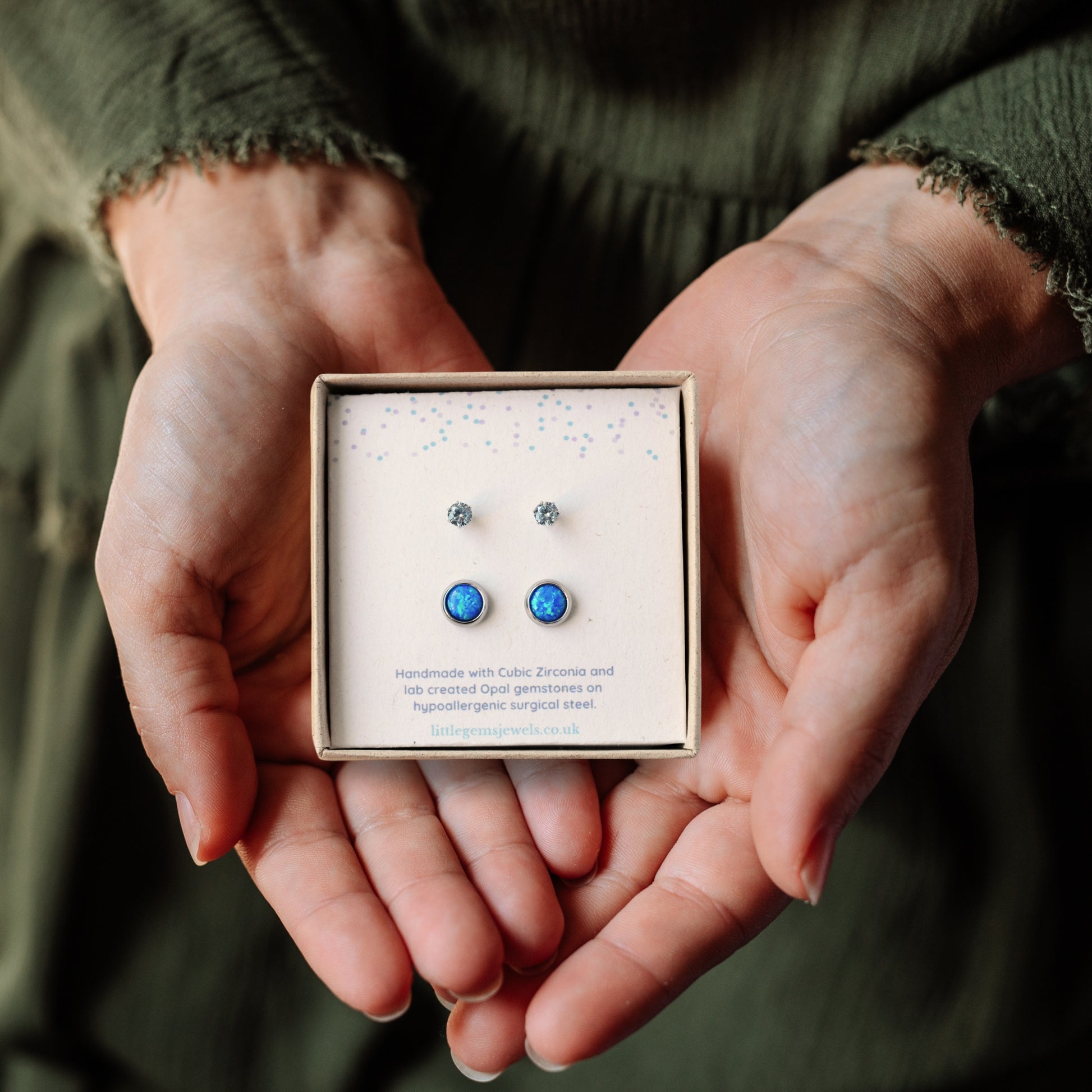 Hands holding a square gift box containing two pairs of blue stone stud earrings.