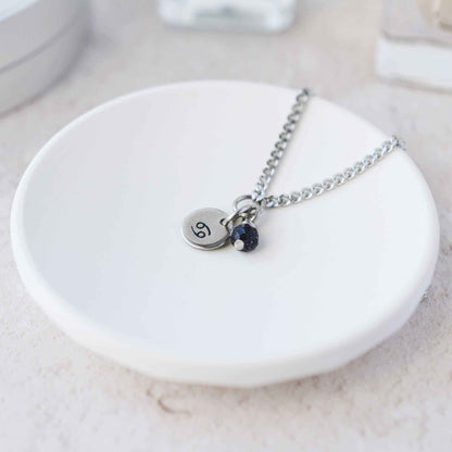 Tiny Cancer zodiac sign disc pendant necklace with Blue Goldstone charm laying on white trinket dish