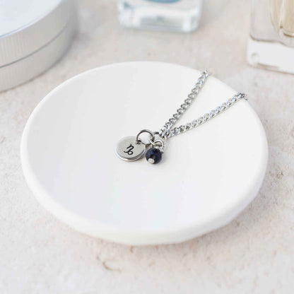 Tiny Capricorn zodiac sign disc pendant necklace with Blue Goldstone charm laying on white trinket dish