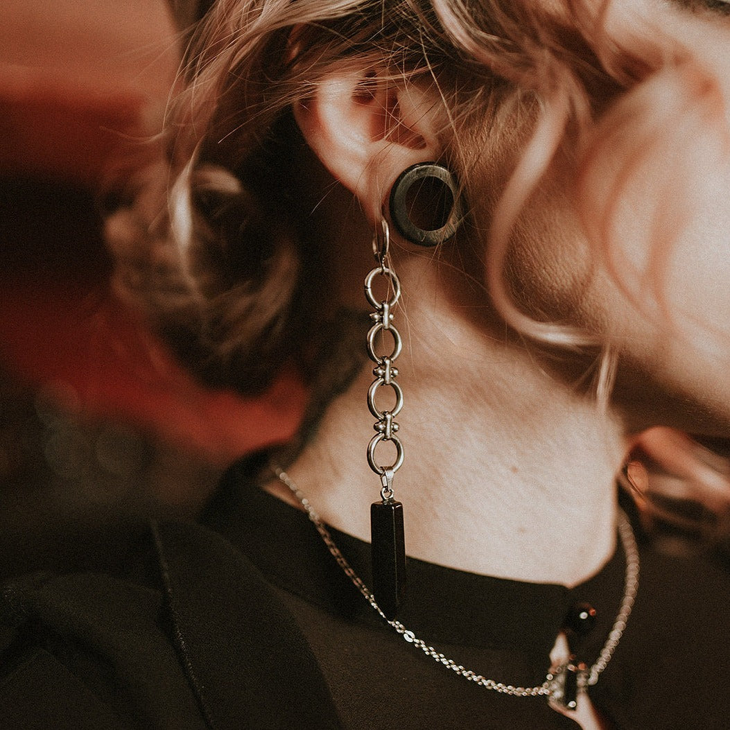Close-up of a person wearing a unique earring with a chain and black pendant.