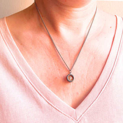 A close-up of a person's neck and chest wearing a silver-toned chain necklace with a circular pendant featuring a yellow gemstone in the center, paired with a light pink V-neck top.