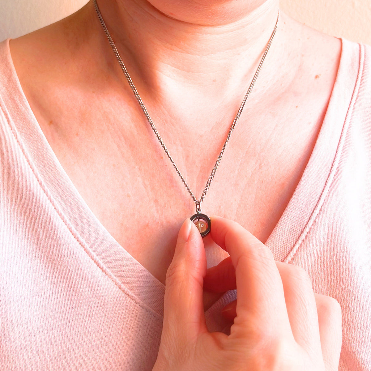 A close-up of a person wearing a silver chain necklace with a circular pendant, touching the pendant with their fingers. The person is wearing a light pink V-neck top against a soft beige background.