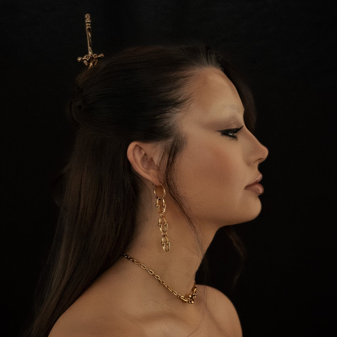 Woman wearing gold jewelry including an earring and necklace against a dark background
