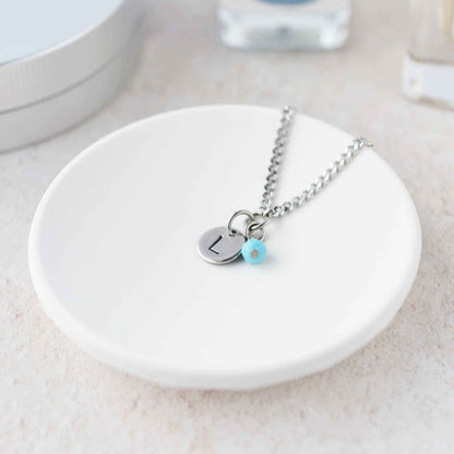 Tiny necklace with round disc pendant stamped with letter L and Turquoise gemstone charm laying in a white trinket dish